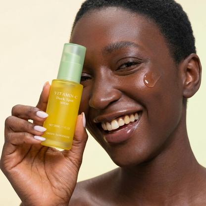 Woman holding a skincare bottle with a droplet of product on her cheek against a beige background